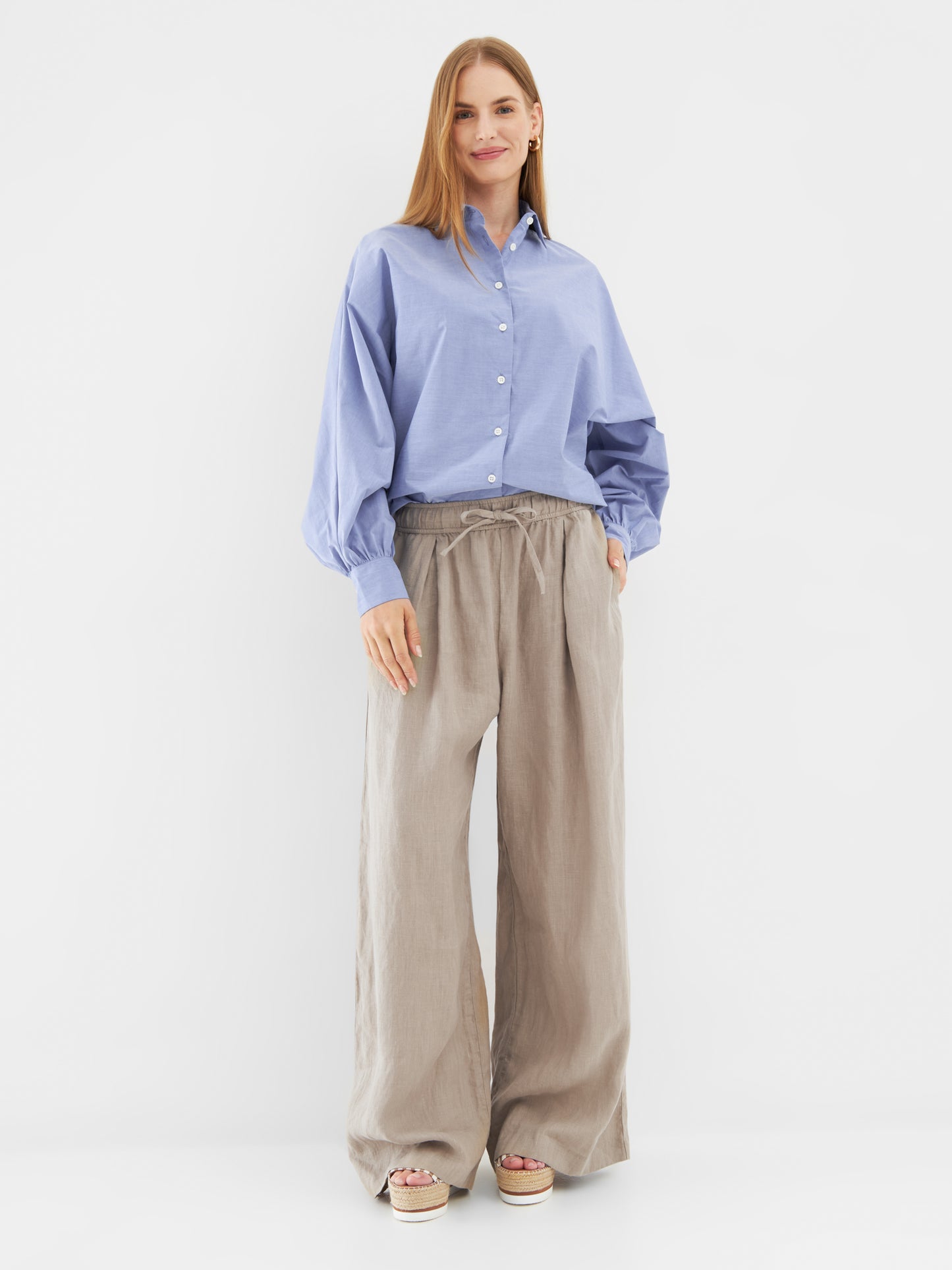 Eine Frau steht vor einem weißen Hintergrund und trägt ein Bluse Antonia Slim Middleblue mit Ballonärmeln, eine beigefarbene Hose mit weitem Bein und Plateausandalen in Beige. Sie hat glattes hellbraunes Haar und ein leichtes Lächeln.