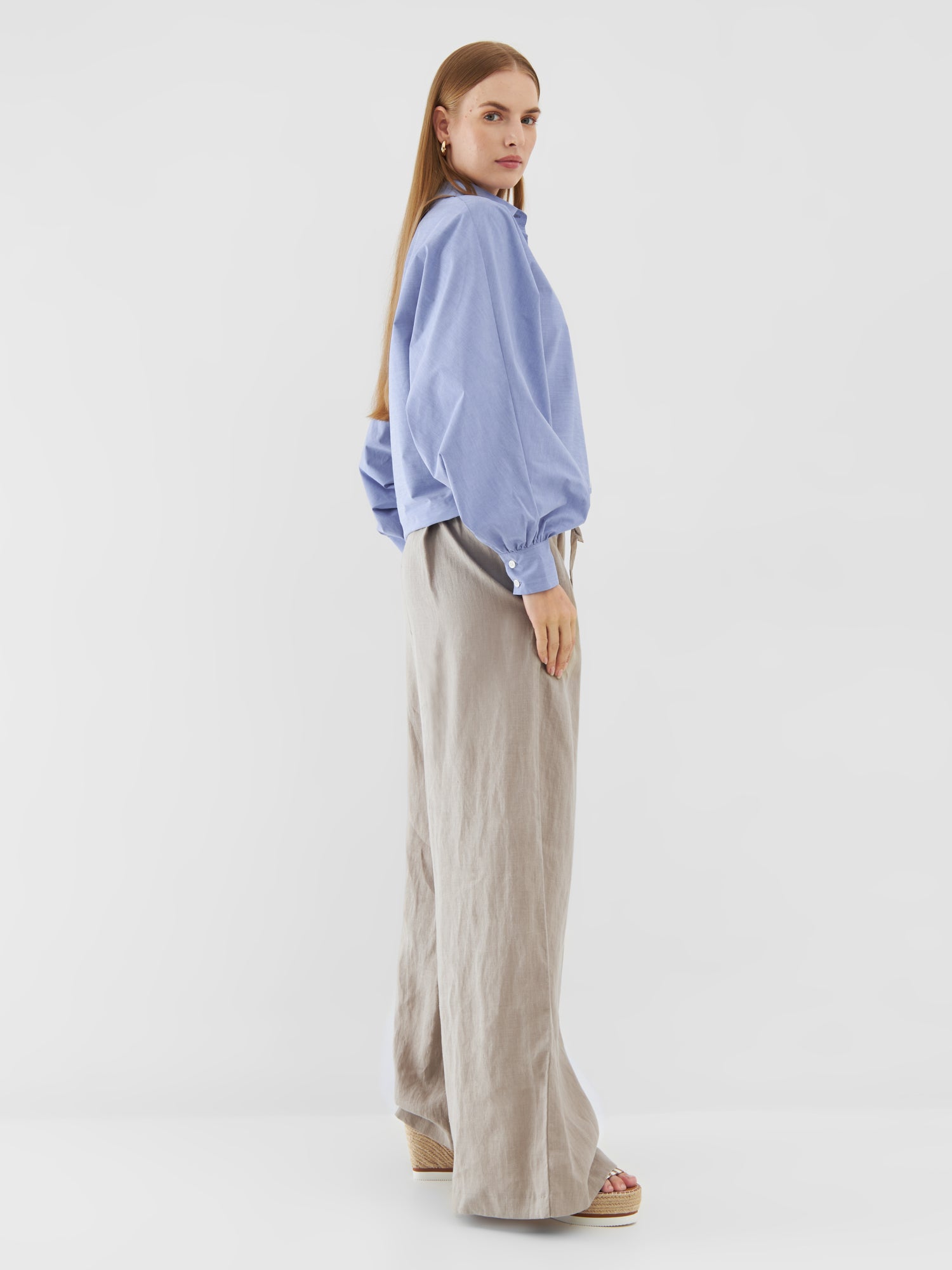 Eine Frau mit langem glattem Haar und neutralem Gesichtsausdruck steht seitlich vor einem weißen Hintergrund. Sie trägt die Bluse Antonia Slim Middleblue, eine beigefarbene Hose mit weitem Bein und Keilsandalen.
