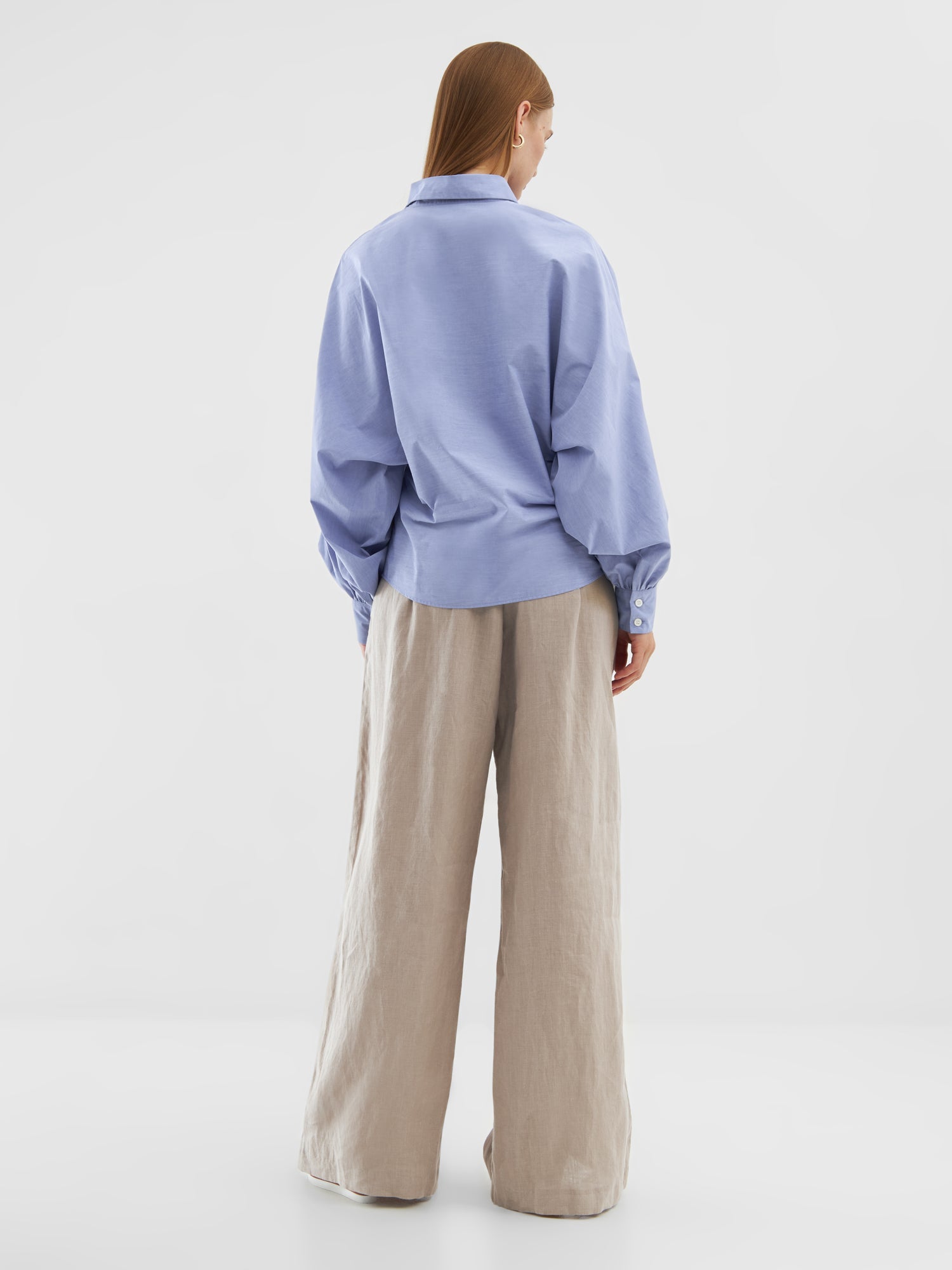 Eine Person mit glattem, langem Haar steht in einer Bluse Antonia Slim Middleblue und einer weitbeinigen beigen Hose vor einem schlichten weißen Hintergrund.