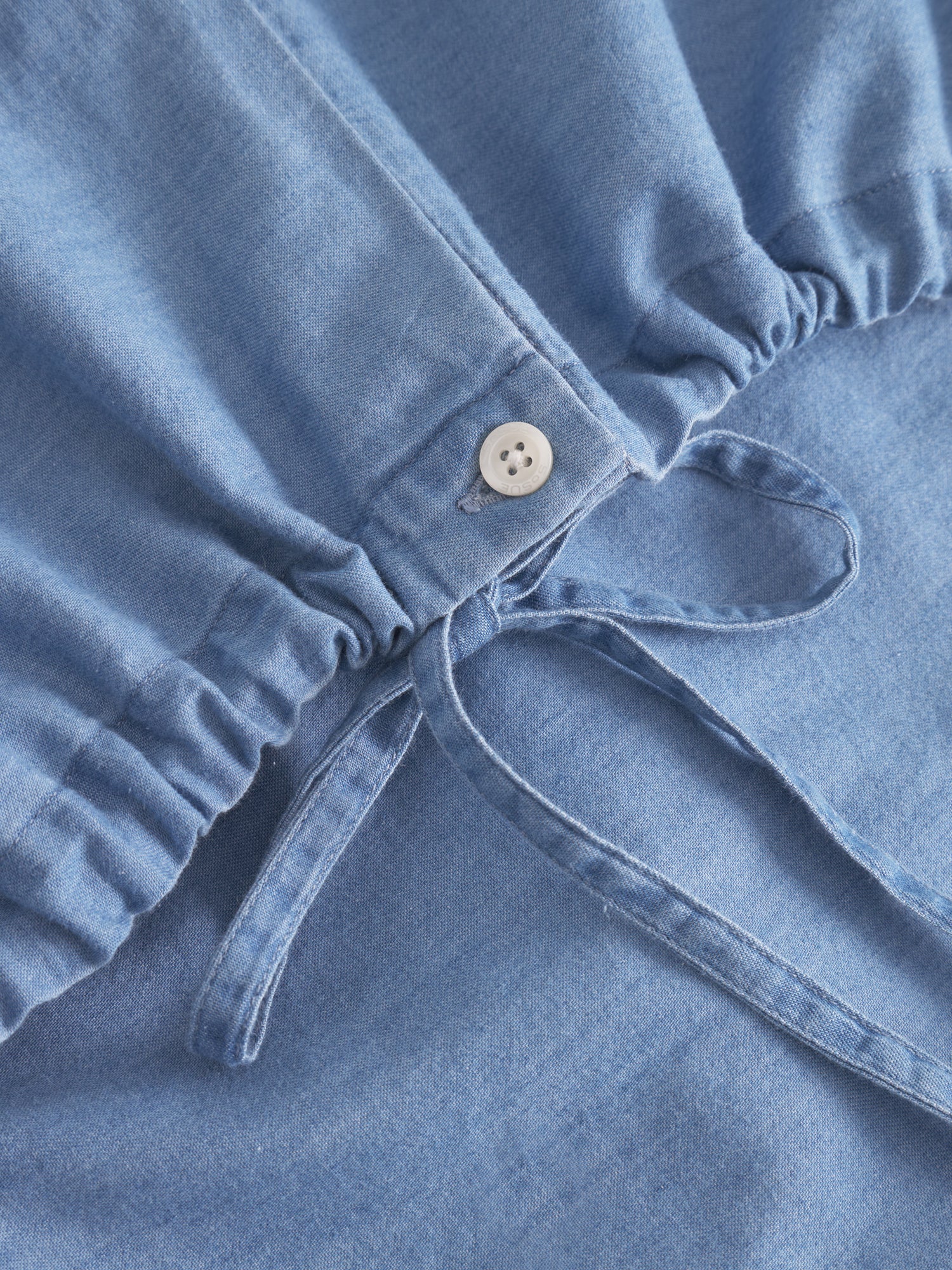 Nahaufnahme der Bluse Antonia Drawstring Denim mit elastischem Bund, weißem Knopf und Kordelzug. Die weiche Textur und das geraffte Taillendetail sorgen für einen stilvollen Look in Denimoptik.
