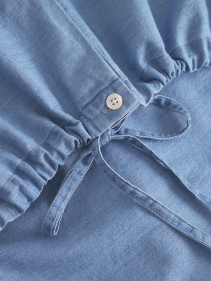 Nahaufnahme der Bluse Antonia Drawstring Denim mit elastischem Bund, weißem Knopf und Kordelzug. Die weiche Textur und das geraffte Taillendetail sorgen für einen stilvollen Look in Denimoptik.