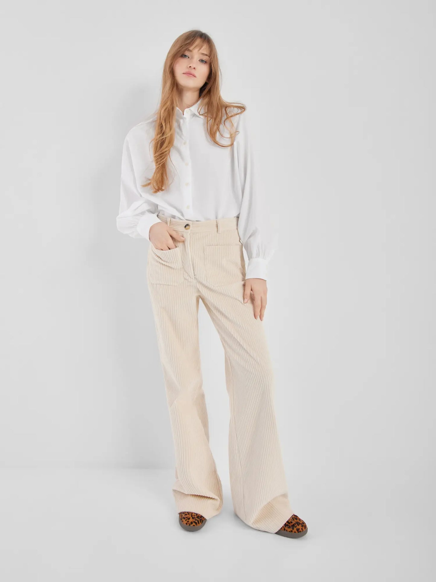 Eine Frau steht vor einem schlichten Hintergrund und trägt die Bluse Antonia Slim White, die in eine hochtaillierte cremefarbene Cordhose gesteckt ist, mit Schuhen mit Leopardenmuster, ihr langes Haar ist offen und ihr Gesichtsausdruck entspannt.