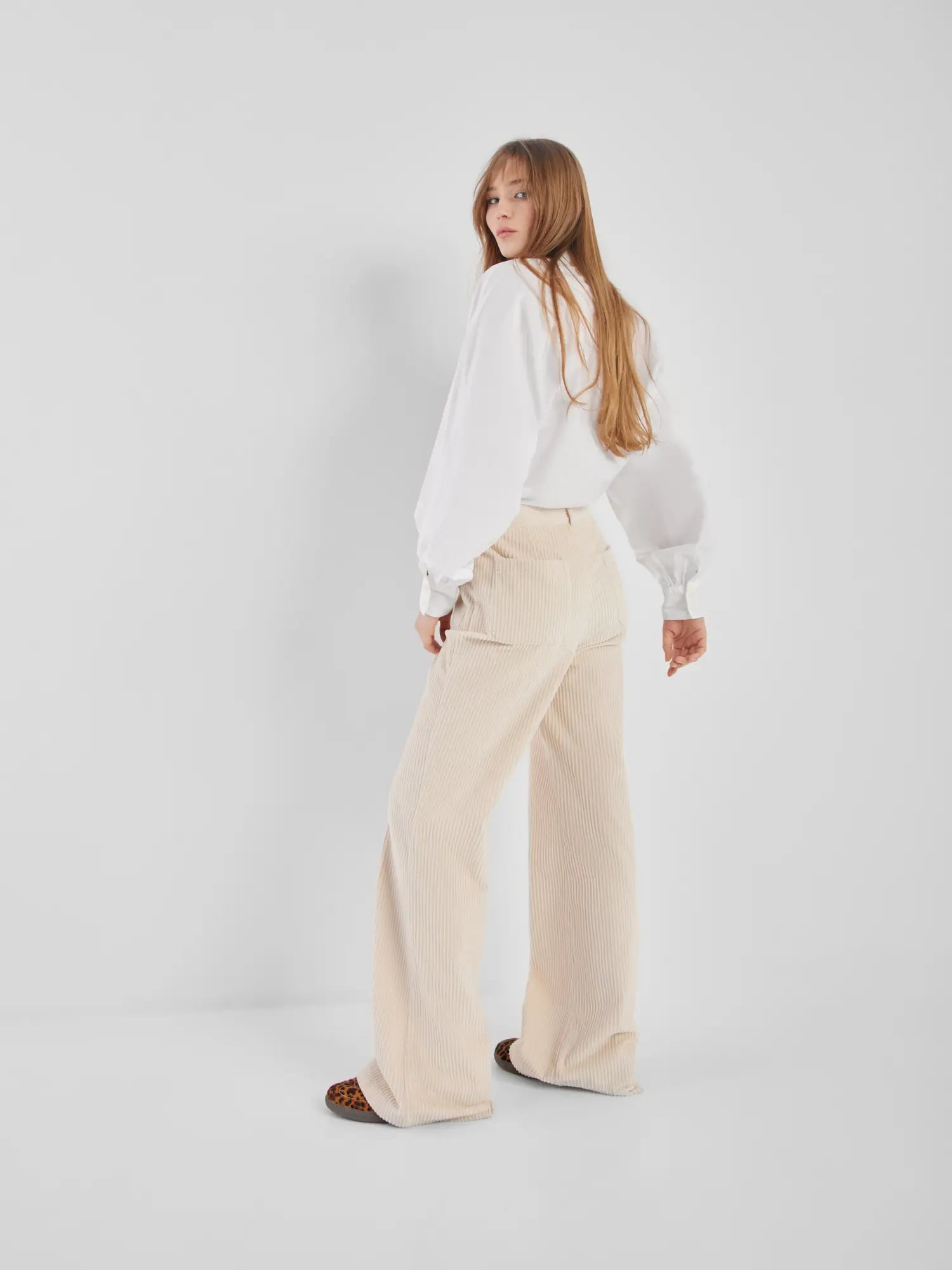 Eine Frau mit langen Haaren trägt eine Bluse Antonia Slim White, eine beigefarbene, weit geschnittene Cordhose und Schuhe mit Leopardenmuster. Sie steht vor einem schlichten weißen Hintergrund und blickt über ihre Schulter.
