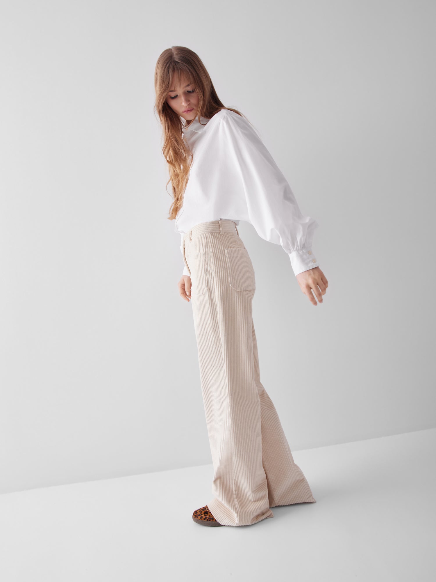 Eine Person mit langen Haaren trägt die Bluse Antonia Slim White und eine beigefarbene Hose mit hoher Taille und weitem Bein. Sie posiert vor einem schlichten hellen Hintergrund und blickt mit einem leicht ausgestreckten Arm nach unten.
