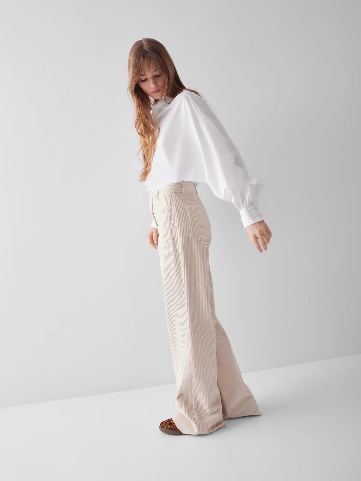 Eine Frau mit langen Haaren trägt eine weiße Bluse Antonia Slim mit voluminösen Ärmeln und eine beigefarbene Cordhose mit hoher Taille und weitem Bein und steht in einem minimalistischen weißen Studio.