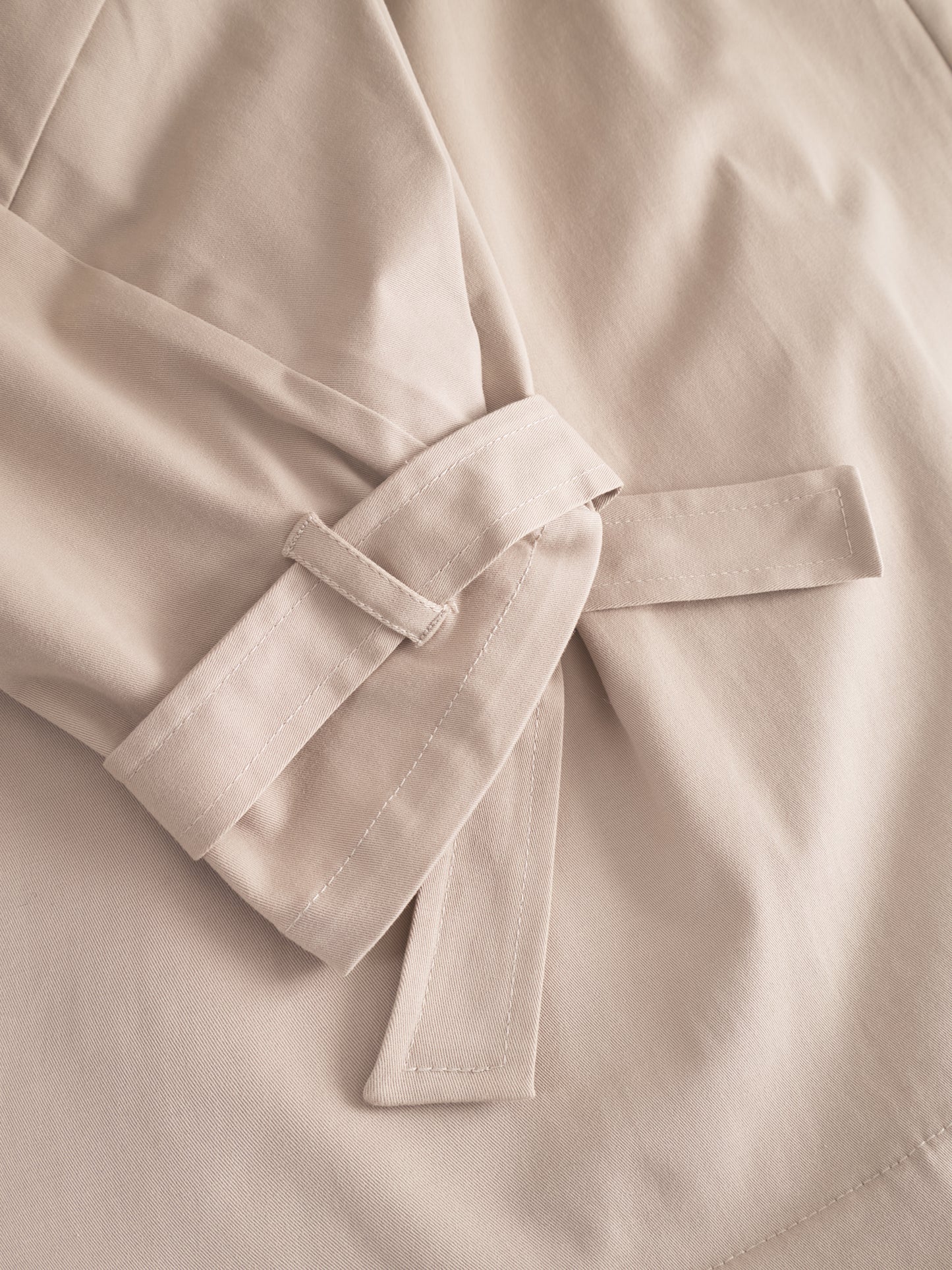 Nahaufnahme eines beigefarbenen Stoffgürtels, der an der Trench Antonia geknotet wird, wobei die detaillierten Nähte und die weiche, glatte Textur hervorgehoben werden.