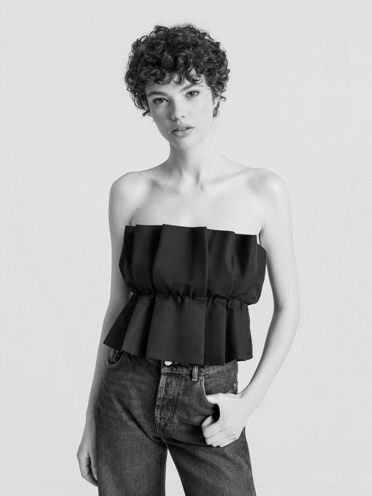 Eine Person mit kurzen Locken trägt das trägerlose „Pleat Top Black“ mit Plisseefalten und eine hoch taillierte Jeans und steht vor einem schlichten Hintergrund, wobei sie eine Hand in die Tasche gesteckt hat. Das Schwarz-Weiß-Bild hebt das Outfit besonders hervor.