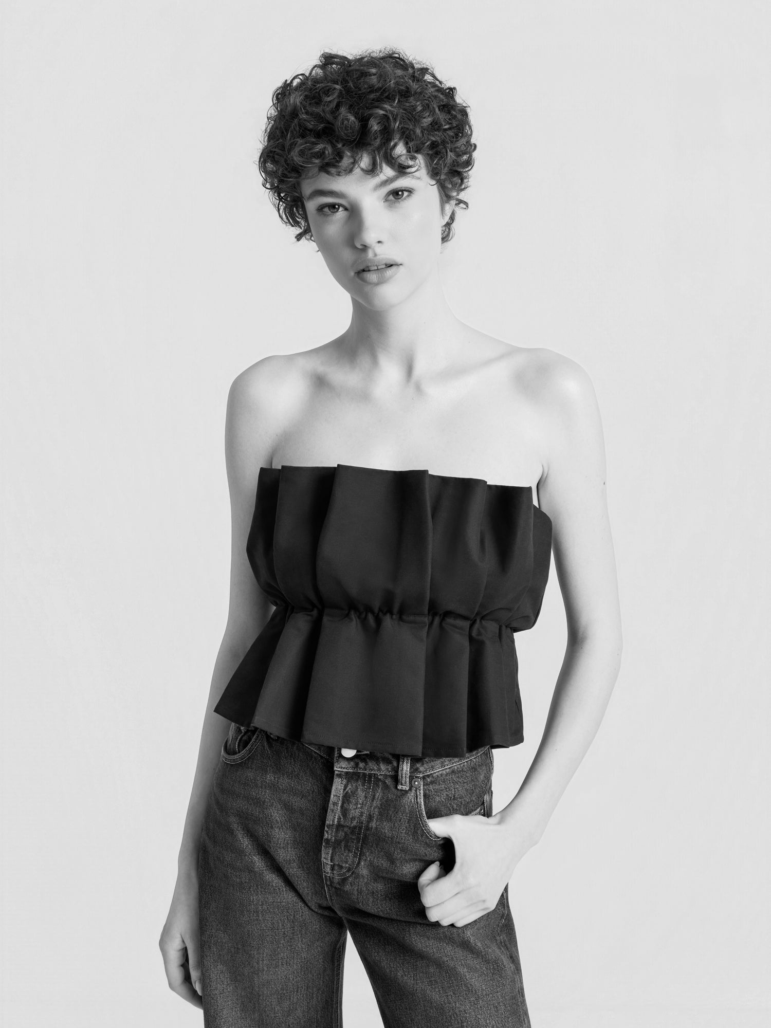 Eine Person mit kurzen Locken trägt das „Pleat Top Black“ – ein trägerloses, plissiertes schwarzes Oberteil – und eine Jeans mit hoher Taille und steht vor einem hellen, einfarbigen Hintergrund, wobei sie eine Hand in die Tasche gesteckt hat.