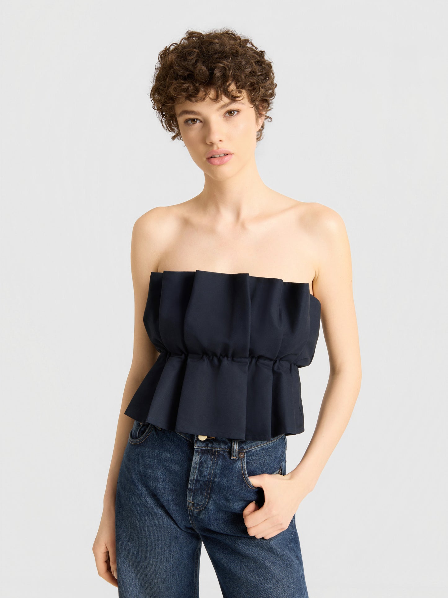 Eine Person mit kurzen, lockigen Haaren trägt ein schwarzes „Pleat Top“ und eine blaue Jeans und steht in einer entspannten Haltung und mit neutralem Gesichtsausdruck vor einem schlichten hellgrauen Hintergrund.