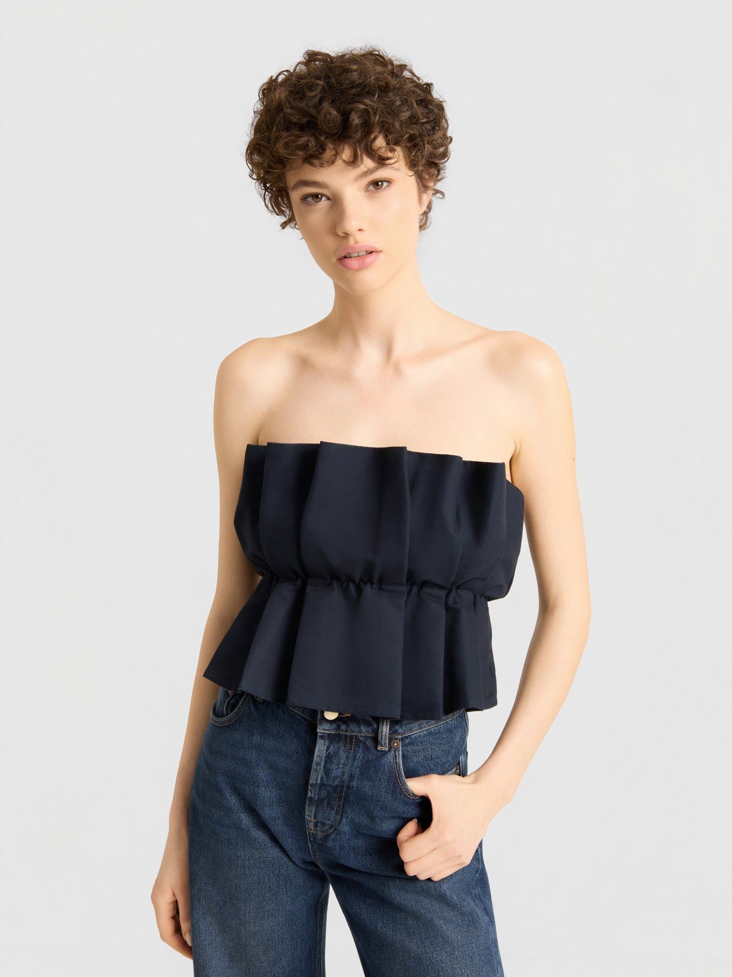 Eine Person mit kurzen, lockigen Haaren trägt ein schwarzes „Pleat Top“ und eine blaue Jeans und steht in einer entspannten Haltung und mit neutralem Gesichtsausdruck vor einem schlichten hellgrauen Hintergrund.