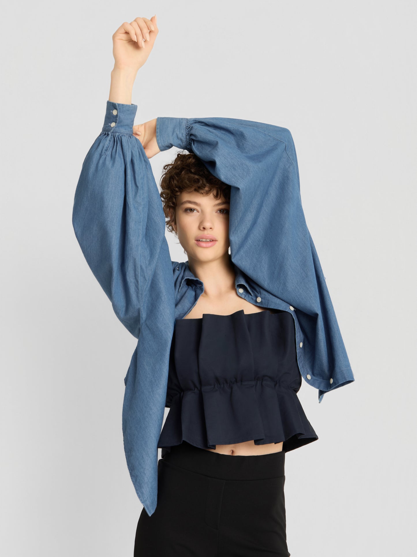 Eine Person mit kurzen Locken posiert mit erhobenem Arm vor einem schlichten hellen Hintergrund. Sie trägt ein blaues Jeanshemd mit übergroßen Ärmeln über einem schwarzen „Pleat Top“ sowie eine schwarze Hose mit hoher Taille.
