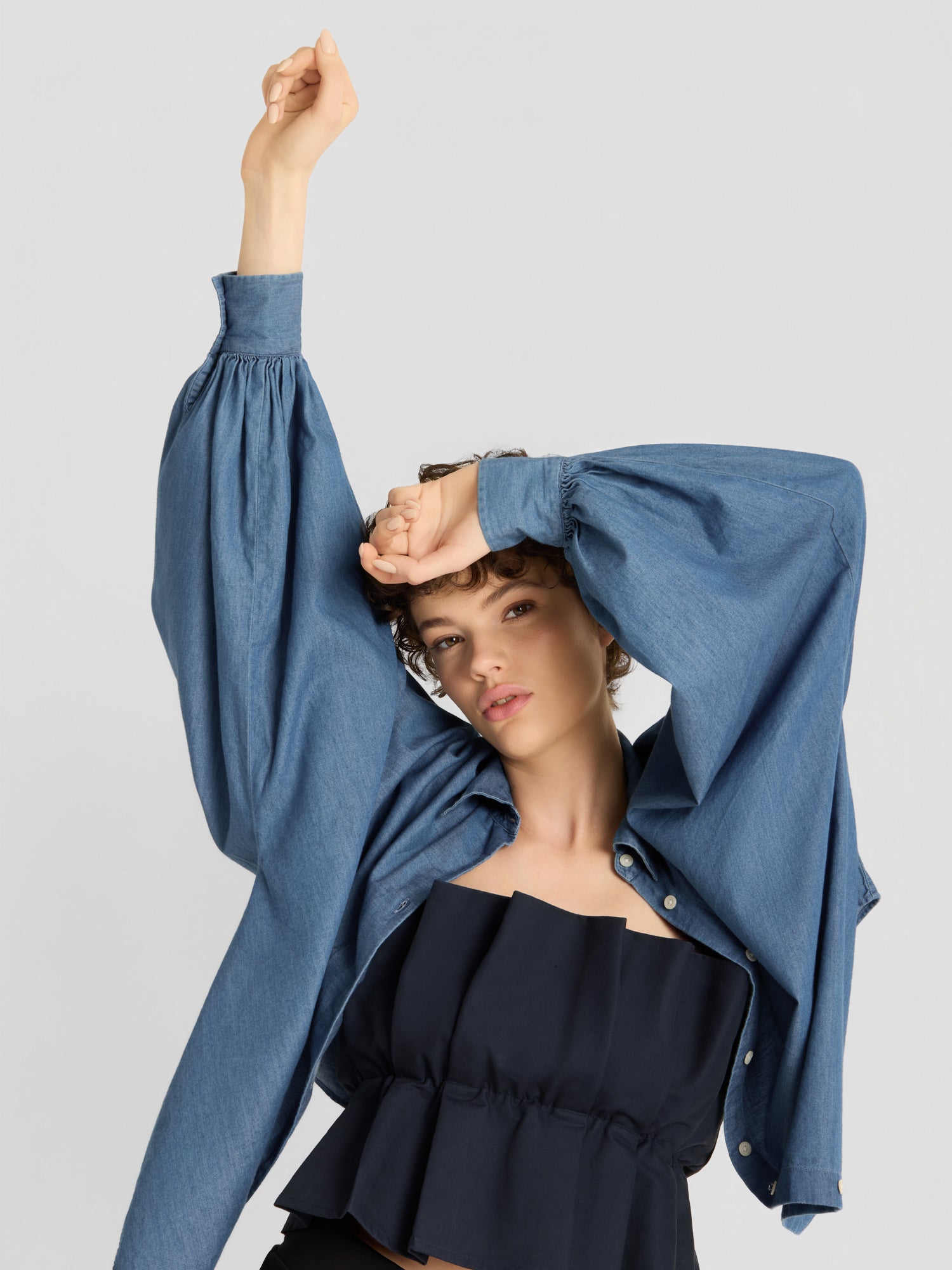 Eine Person, die ein lockeres blaues Jeanshemd über dem „Pleat Top Black“ trägt, posiert mit einem erhobenen Arm und dem anderen Arm angewinkelt über der Stirn vor einem schlichten hellgrauen Hintergrund.