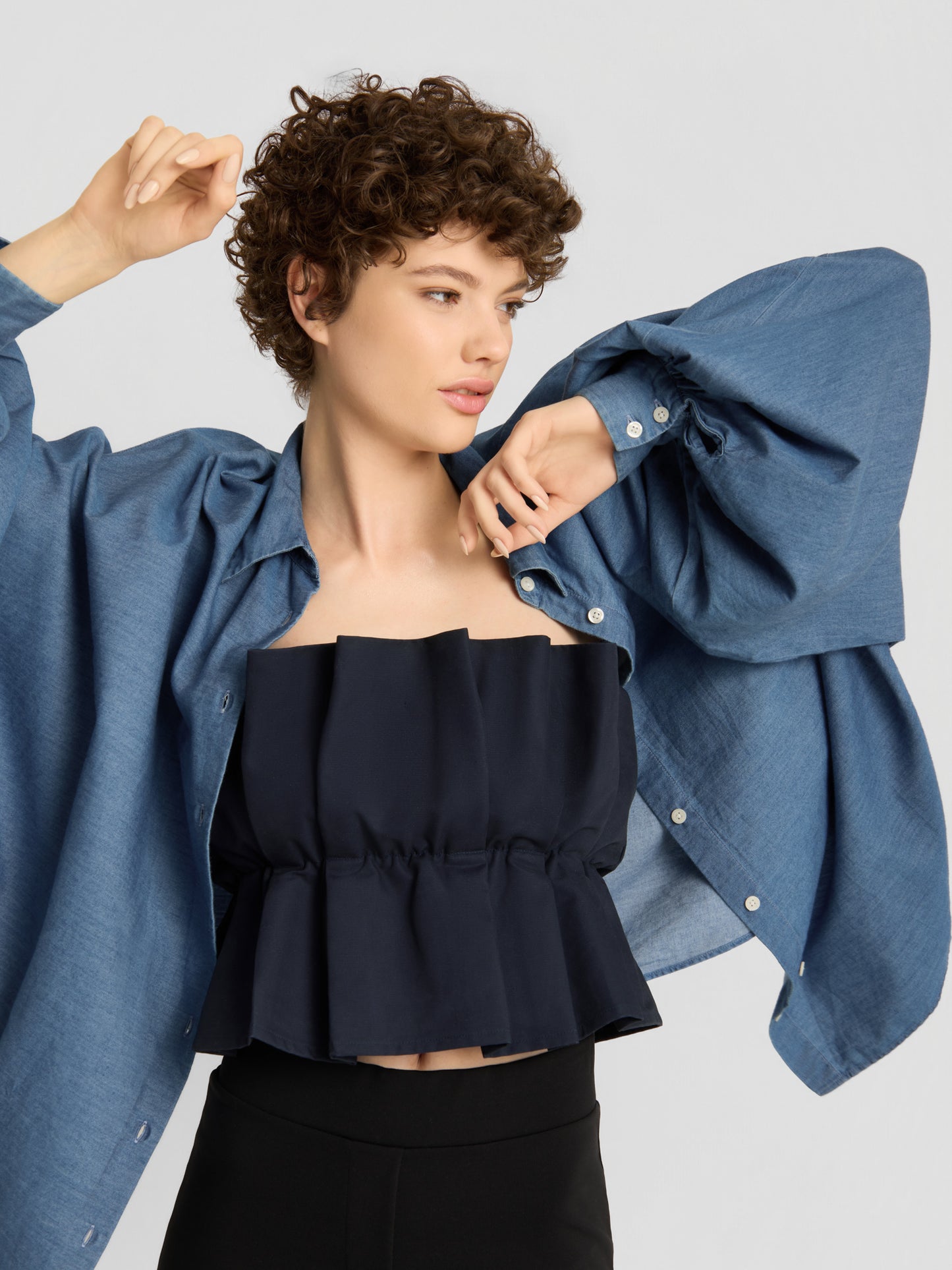 Eine Frau mit kurzen Locken posiert mit erhobenem Arm; sie trägt das „Pleat Top Black“ unter einem lockeren blauen Hemd mit Knöpfen und einer schwarzen Hose vor einem schlichten hellen Hintergrund.