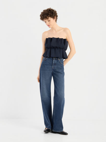 Eine Frau mit kurzen Locken trägt ein schwarzes, plissiertes Oberteil mit Rüschen und eine blaue Jeans mit hoher Taille und weitem Bein. Sie steht vor einem schlichten weißen Hintergrund, blickt nach unten und hat eine Hand in der Tasche.