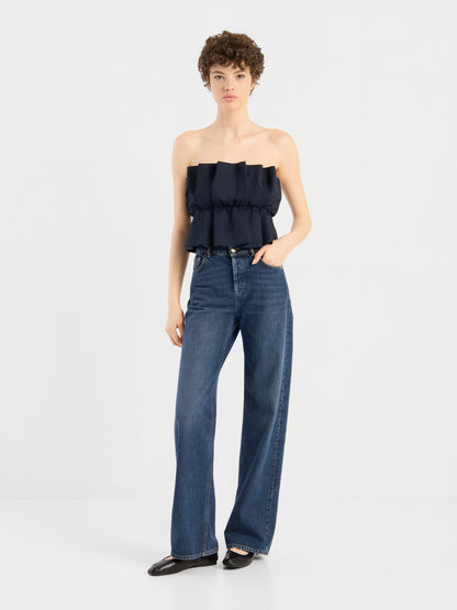 Eine Person mit kurzen Locken trägt ein schwarzes „Pleat Top“ mit Rüschen, dazu eine hoch taillierte, weit geschnittene blaue Jeans und schwarze Schuhe; sie steht vor einem schlichten weißen Hintergrund.