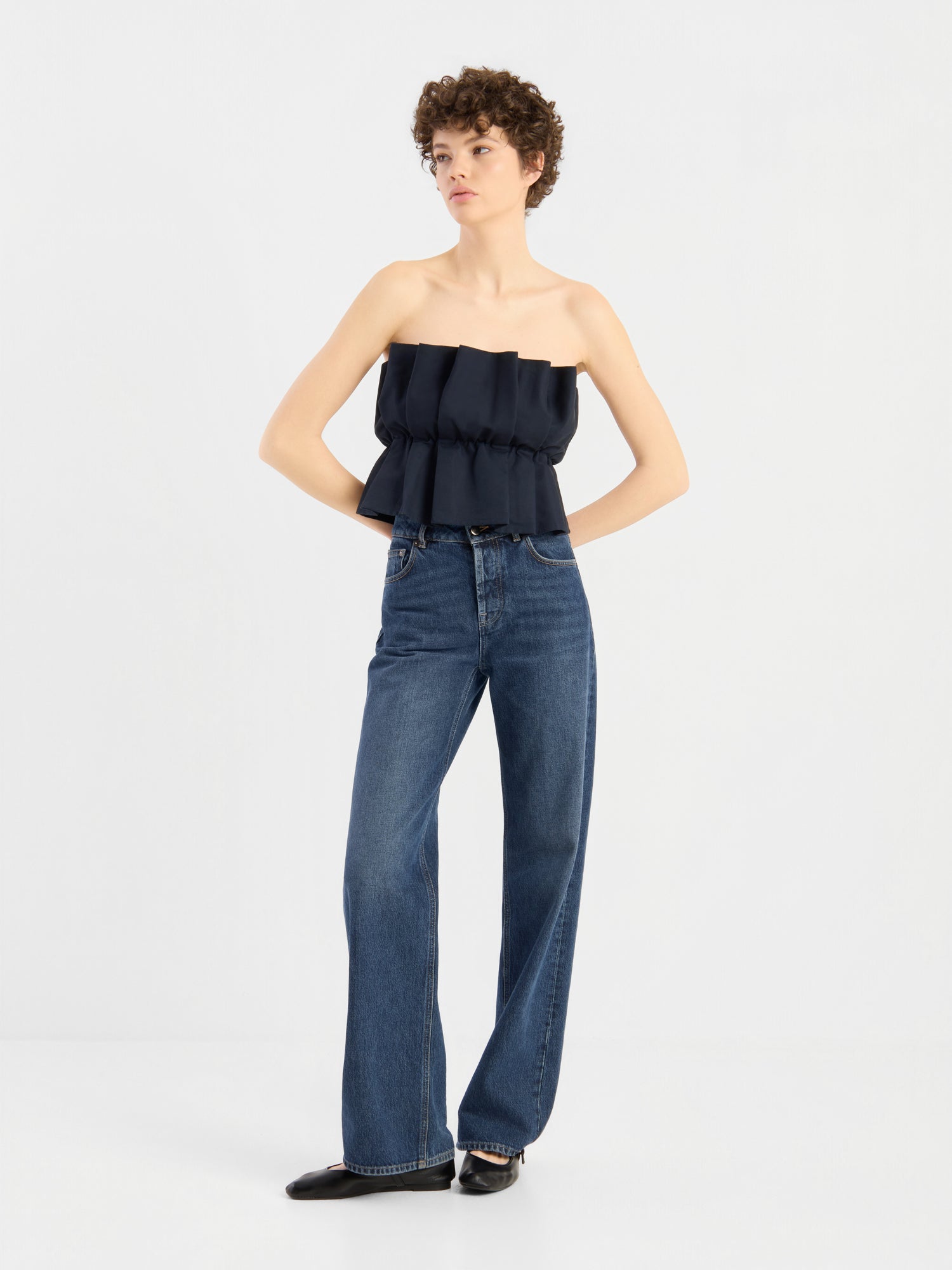 Eine Person mit kurzen Locken trägt ein schwarzes „Pleat Top“, das über einer hoch taillierten, weit geschnittenen blauen Jeans liegt. Sie steht mit den Händen in den Hüften da und blickt vor einem schlichten weißen Hintergrund zur Seite.