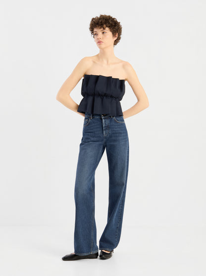 Eine Person mit kurzen Locken trägt ein schwarzes „Pleat Top“, das über einer hoch taillierten, weit geschnittenen blauen Jeans liegt. Sie steht mit den Händen in den Hüften da und blickt vor einem schlichten weißen Hintergrund zur Seite.