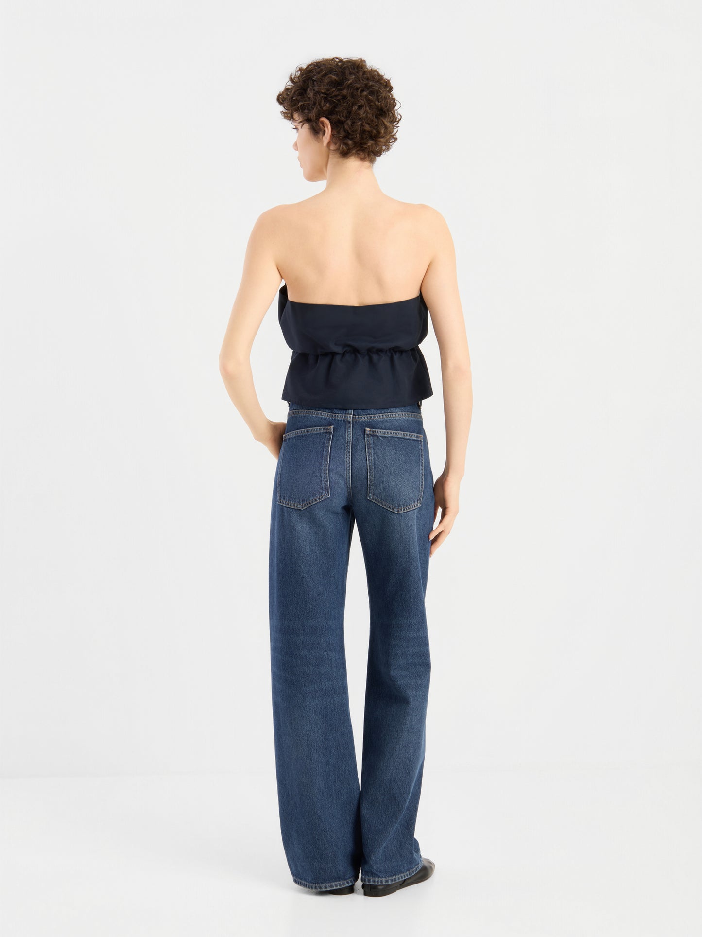 Eine Person mit kurzen Locken, von hinten gesehen, trägt das „Pleat Top Black“ und eine blaue Jeans mit hoher Taille und weitem Bein und steht vor einem schlichten weißen Hintergrund.