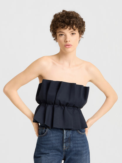 Eine junge Frau mit kurzen Locken trägt das „Pleat Top Black“ mit Plissee-Details und eine blaue Jeans und steht mit den Händen in die Hüften gestemmt vor einem schlichten, hellen Hintergrund.