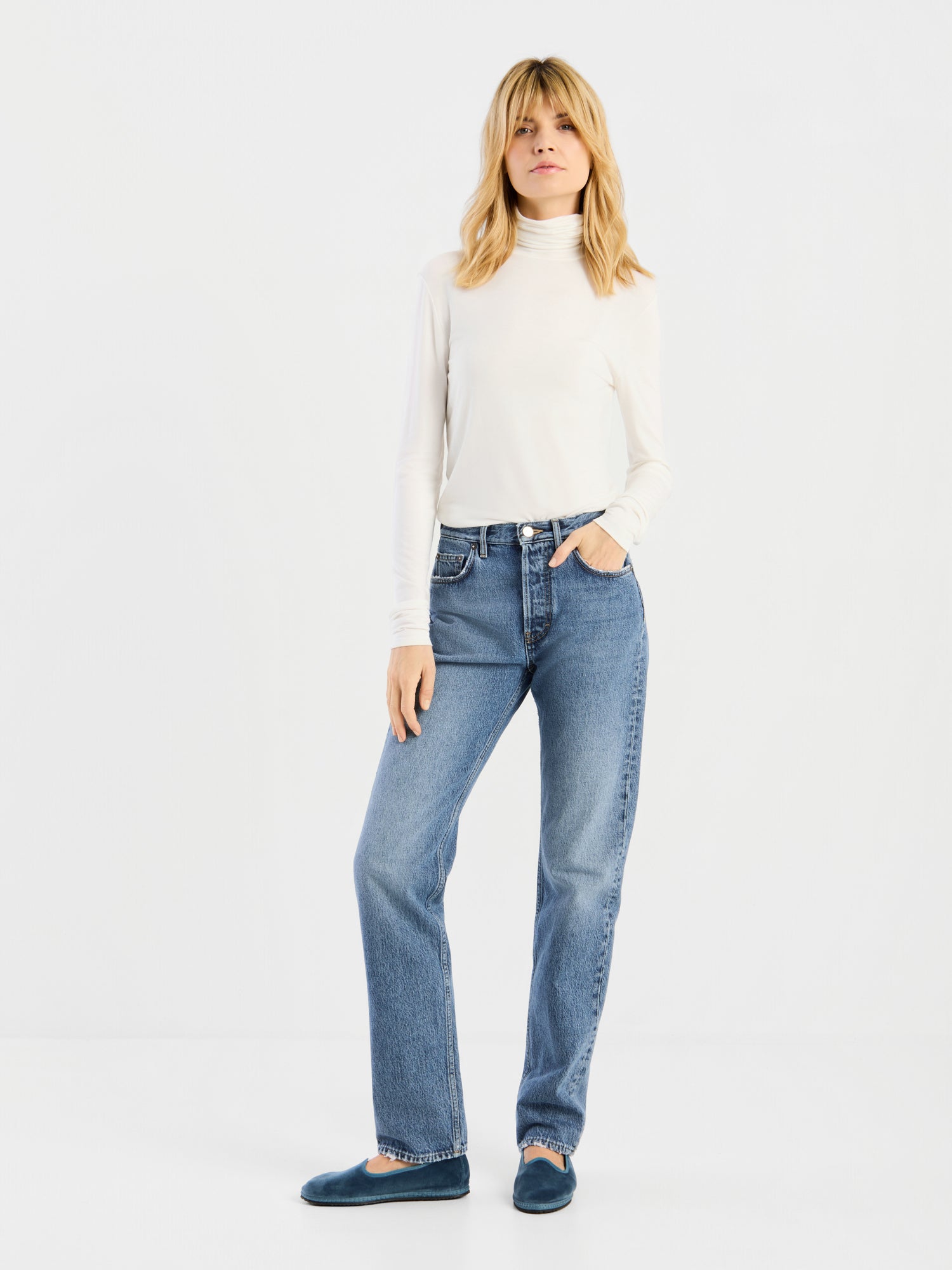 Eine Person steht vor einem schlichten weißen Hintergrund und trägt das „Turtle Jersey White“ in Kombination mit blauen Jeans und dunklen Schuhen; eine Hand steckt in der Hosentasche, und ihr Gesichtsausdruck wirkt entspannt.