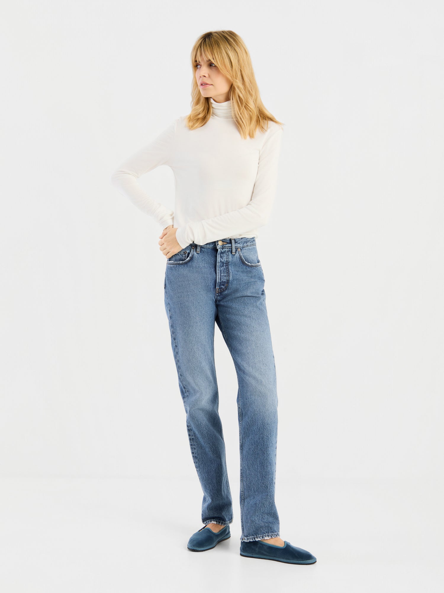 Eine Person mit blonden Haaren trägt das „Turtle Jersey White“, dazu blaue Jeans und dunkelblaue Hausschuhe, und steht vor einem schlichten weißen Hintergrund.