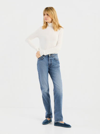 Eine Person mit blonden Haaren trägt das „Turtle Jersey White“, dazu blaue Jeans und dunkelblaue Hausschuhe, und steht vor einem schlichten weißen Hintergrund.