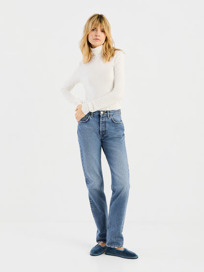 Eine Person, die ein weißes „Turtle“-Trikot, blaue Jeans und blaue Slipper trägt, steht vor einem schlichten weißen Hintergrund und blickt nach vorne, während ihre Hände in Hüfthöhe ruhen.
