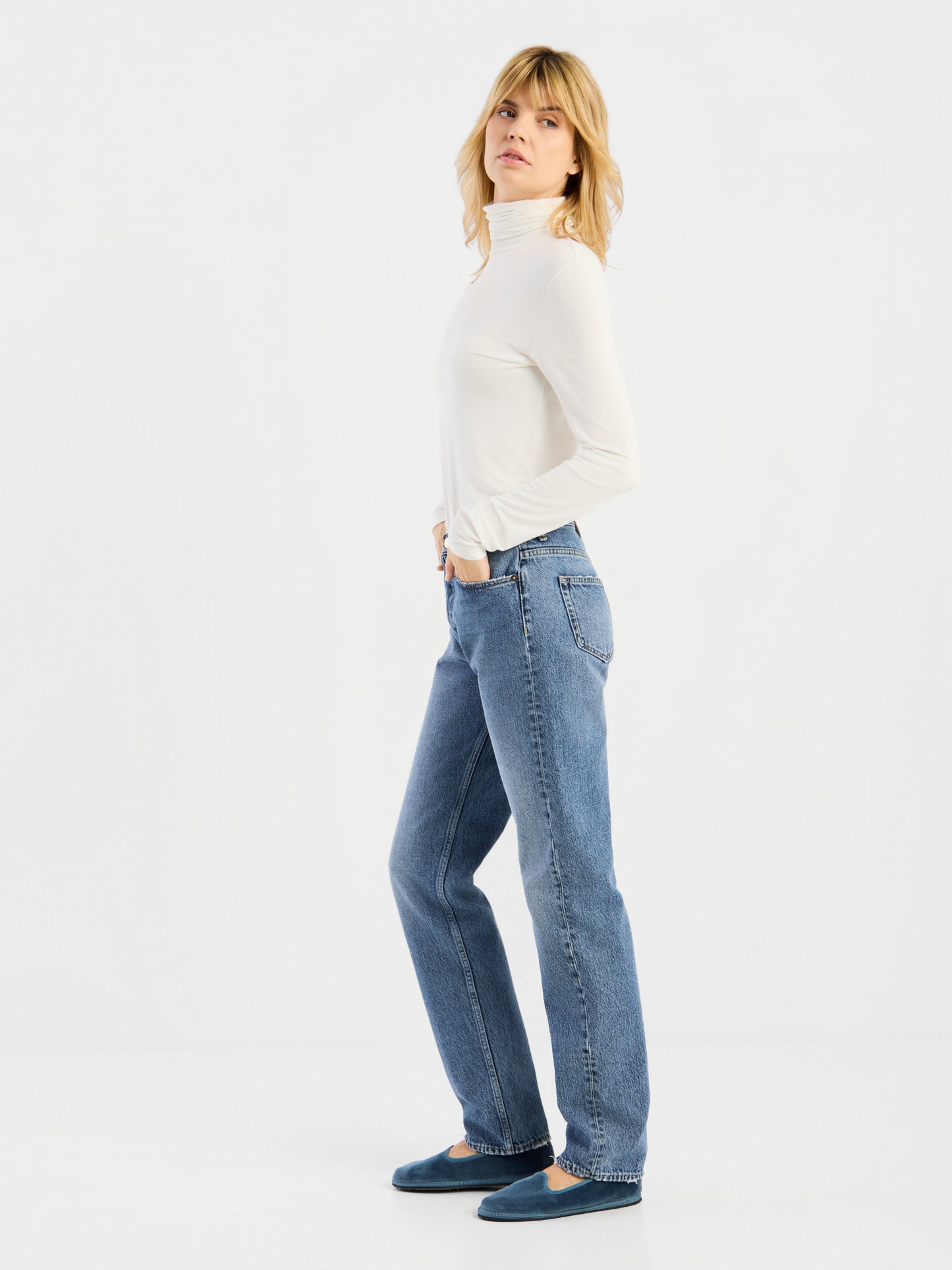 Vor einem schlichten weißen Hintergrund steht eine Person mit blonden Haaren in Seitenansicht und trägt das weiße „Turtle“-Trikot, blaue Jeans und blaue Schuhe.