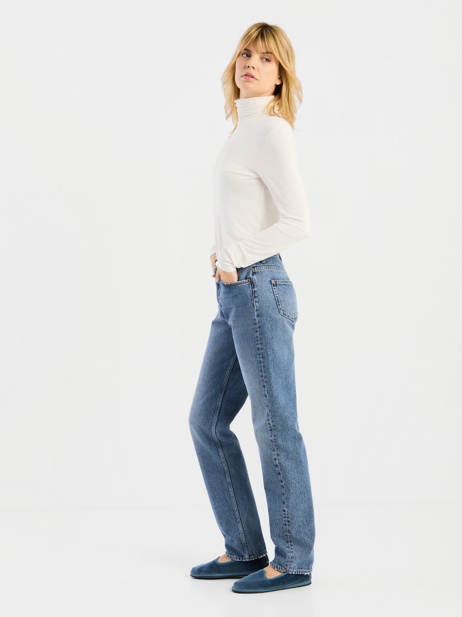 Vor einem schlichten weißen Hintergrund steht eine Person mit blonden Haaren in Seitenansicht und trägt das weiße „Turtle“-Trikot, blaue Jeans und blaue Schuhe.