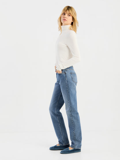 Vor einem schlichten weißen Hintergrund steht eine Person mit blonden Haaren in Seitenansicht und trägt das weiße „Turtle“-Trikot, blaue Jeans und blaue Schuhe.