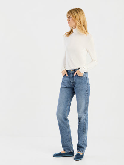 Eine Person steht vor einem schlichten weißen Hintergrund und trägt das „Turtle Jersey White“, blaue Jeans und blaue Ballerinas; die Hände hat sie in den Taschen, den Blick zur Seite gerichtet.