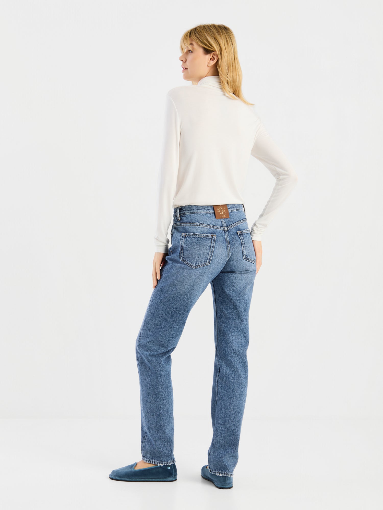 Eine Person steht mit dem Rücken zur Kamera und trägt das langärmelige „Turtle Jersey White“, kombiniert mit einer blauen Straight-Leg-Jeans und blauen Schuhen, vor einem schlichten weißen Hintergrund. Der klassische Look wird durch die erstklassige Qualität des „Turtle Jersey White“ unterstrichen, das stolz in Portugal hergestellt wird.
