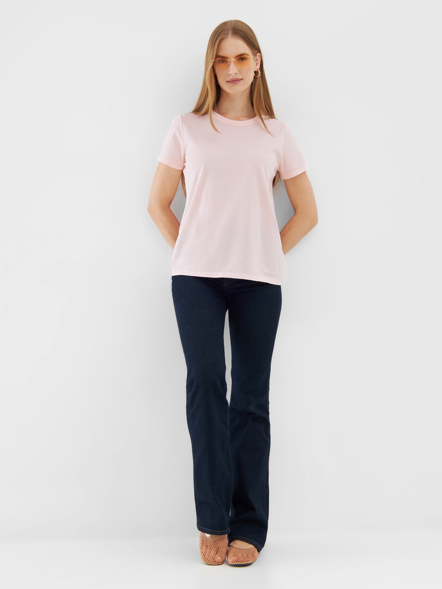 Eine Frau steht vor einem weißen Hintergrund und trägt ein T-Shirt Rose, dunkelblaue Schlagjeans, braune gewebte Schuhe und eine runde Sonnenbrille. Ihre Hände sind hinter ihrem Rücken verschränkt, während sie nach vorne schaut.