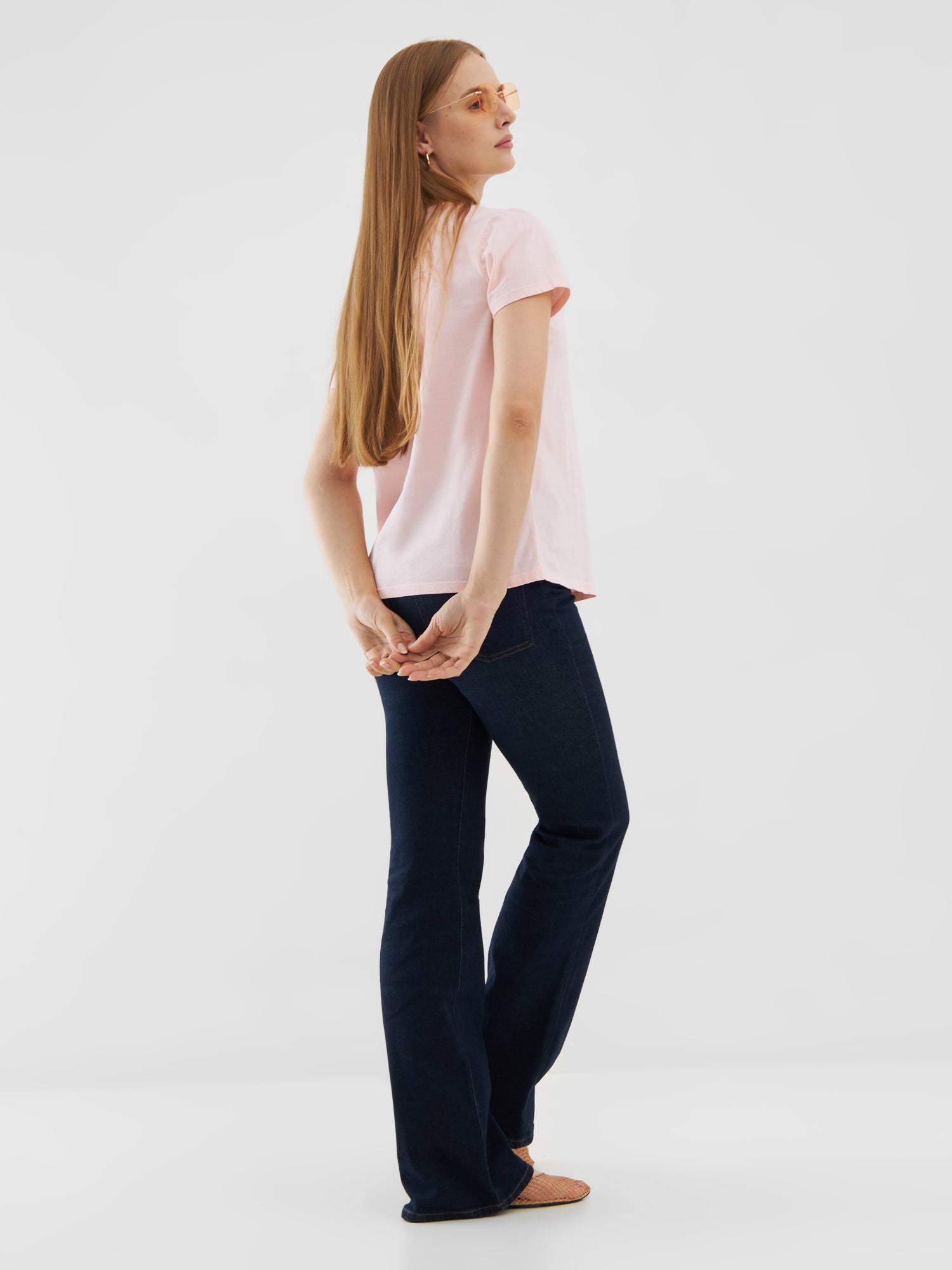 Eine Frau mit langem, glattem Haar, die ein T-Shirt Rose, dunkelblaue Jeans und Sandalen trägt, steht mit dem Blick über die Schulter und mit hinter dem Rücken verschränkten Händen vor einem schlichten weißen Hintergrund.