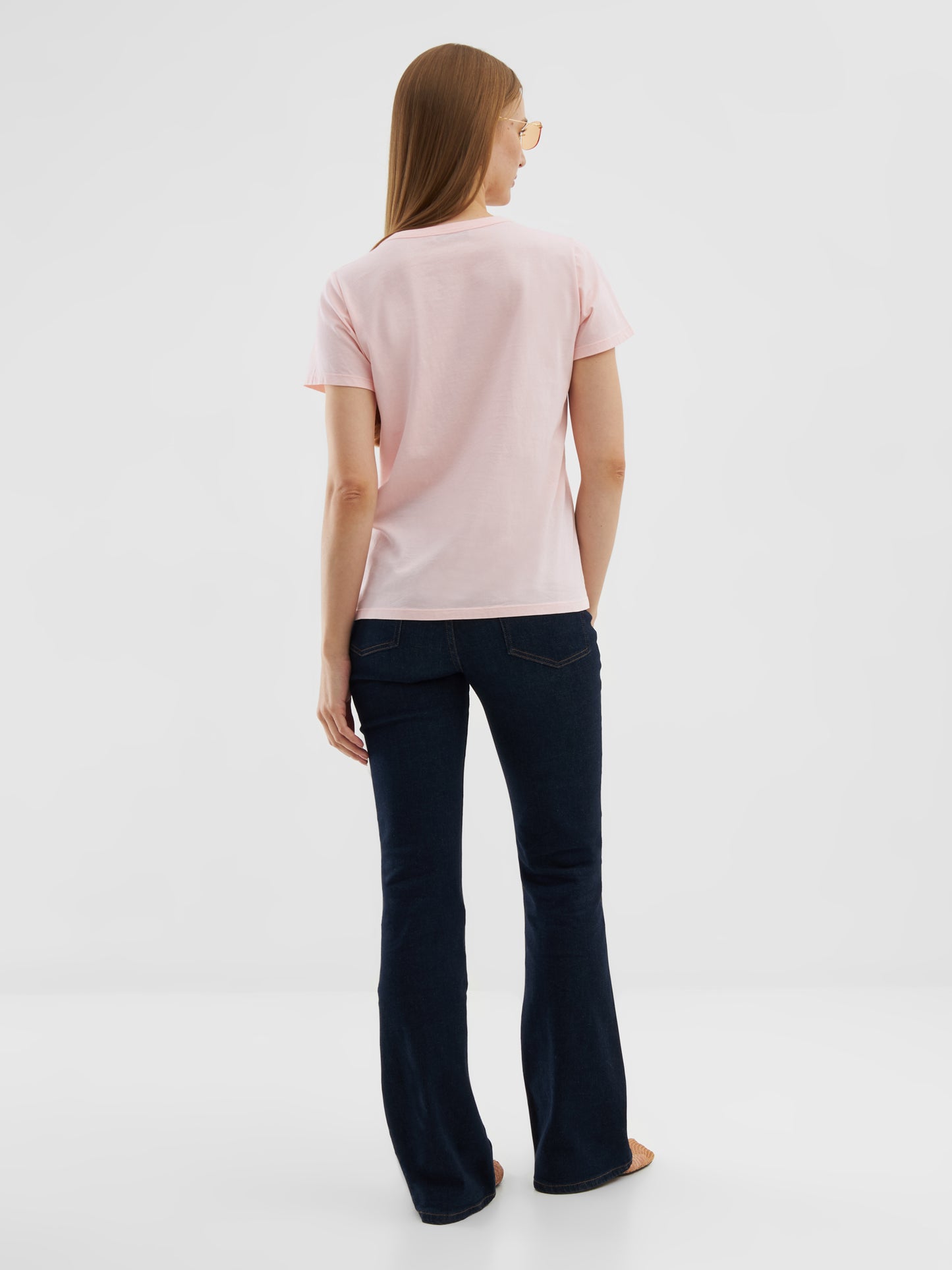 Eine Frau mit glattem, hellbraunem Haar steht vor einem einfarbig weißen Hintergrund. Sie trägt ein T-Shirt Rose aus Baumwolle, dunkelblaue Schlagjeans und eine Sonnenbrille.