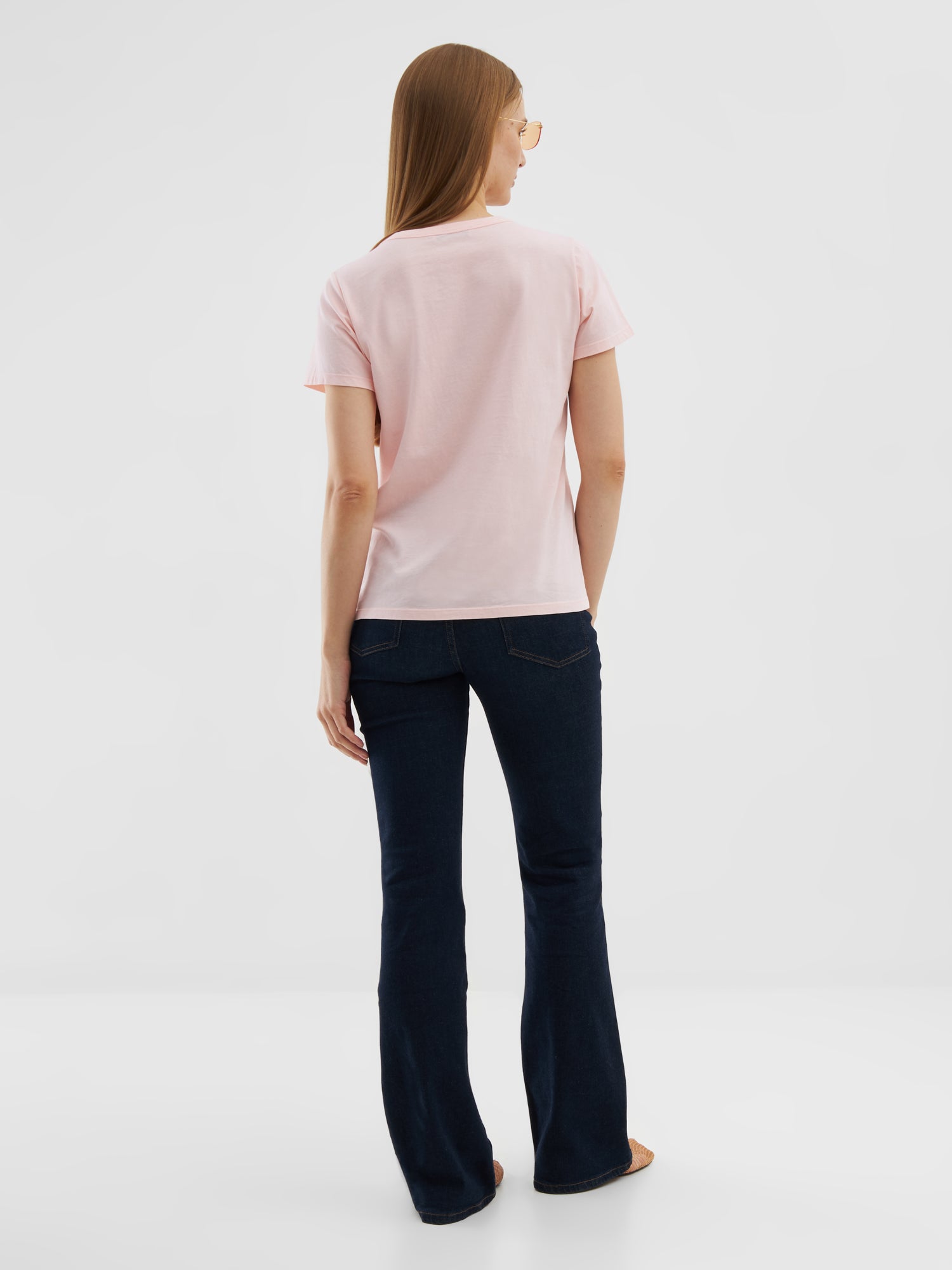 Eine Frau mit glattem, hellbraunem Haar steht vor einem einfarbig weißen Hintergrund. Sie trägt ein T-Shirt Rose aus Baumwolle, dunkelblaue Schlagjeans und eine Sonnenbrille.