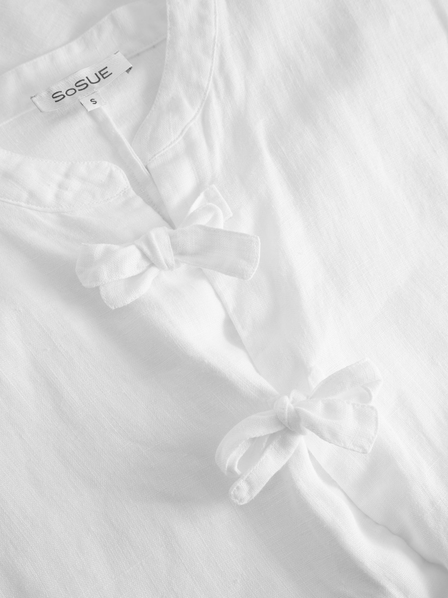 Nahaufnahme der Bluse Cowboy Bow mit sichtbarem SoSUE-Label und zwei Stoffschleifen auf der Vorderseite. Hergestellt aus weichem, strukturiertem Material - ideal für Liebhaber nachhaltiger Mode.