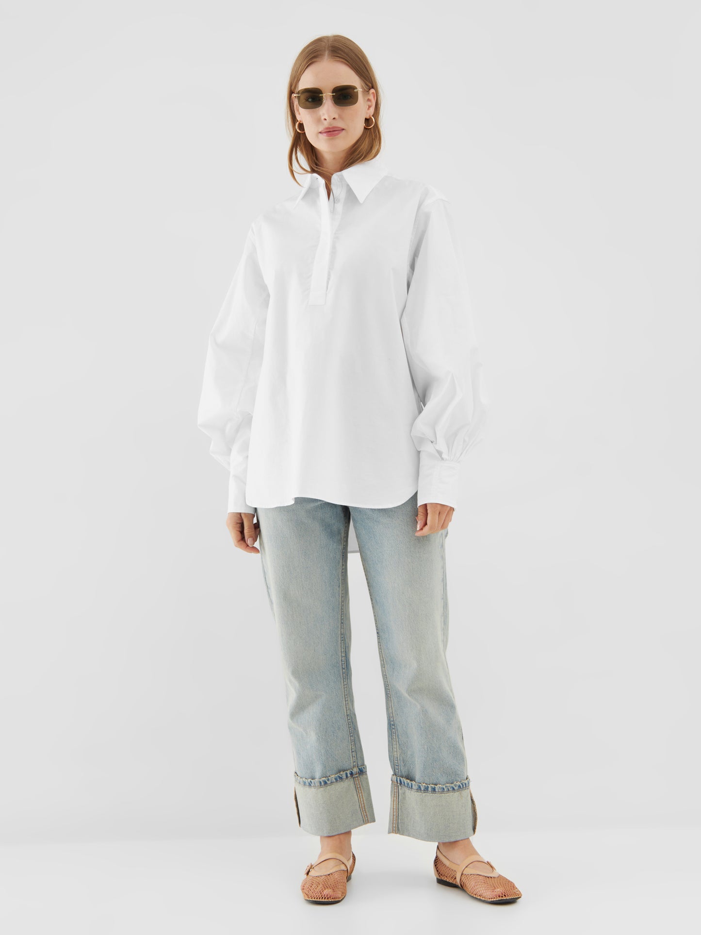 Eine Person mit Sonnenbrille, der übergroßen Bluse Rini White mit Ballonärmeln, hellblauen Jeans mit Bündchen und gewebten hellbraunen Flats steht vor einem schlichten weißen Hintergrund.