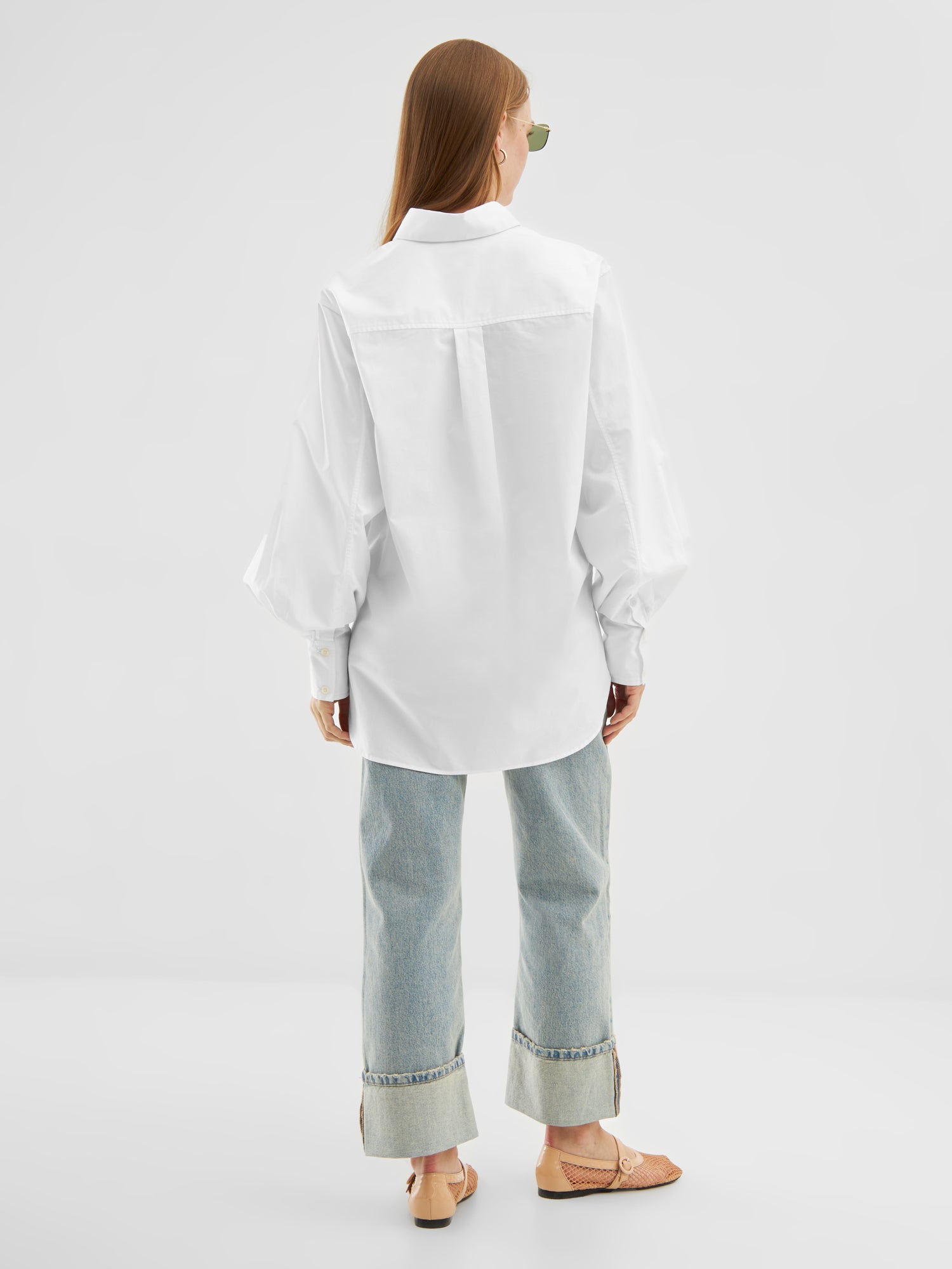 Eine Person mit langen Haaren und Sonnenbrille trägt die Bluse Rini White aus Baumwollpopeline, hellblaue Jeans mit weitem Bein und breiten Bündchen sowie beigefarbene Sandalen und steht mit dem Rücken zu einem einfarbig weißen Hintergrund.