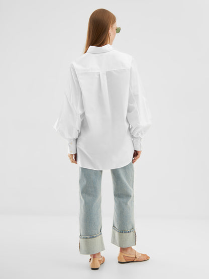 Eine Person mit langen Haaren und Sonnenbrille trägt die Bluse Rini White aus Baumwollpopeline, hellblaue Jeans mit weitem Bein und breiten Bündchen sowie beigefarbene Sandalen und steht mit dem Rücken zu einem einfarbig weißen Hintergrund.