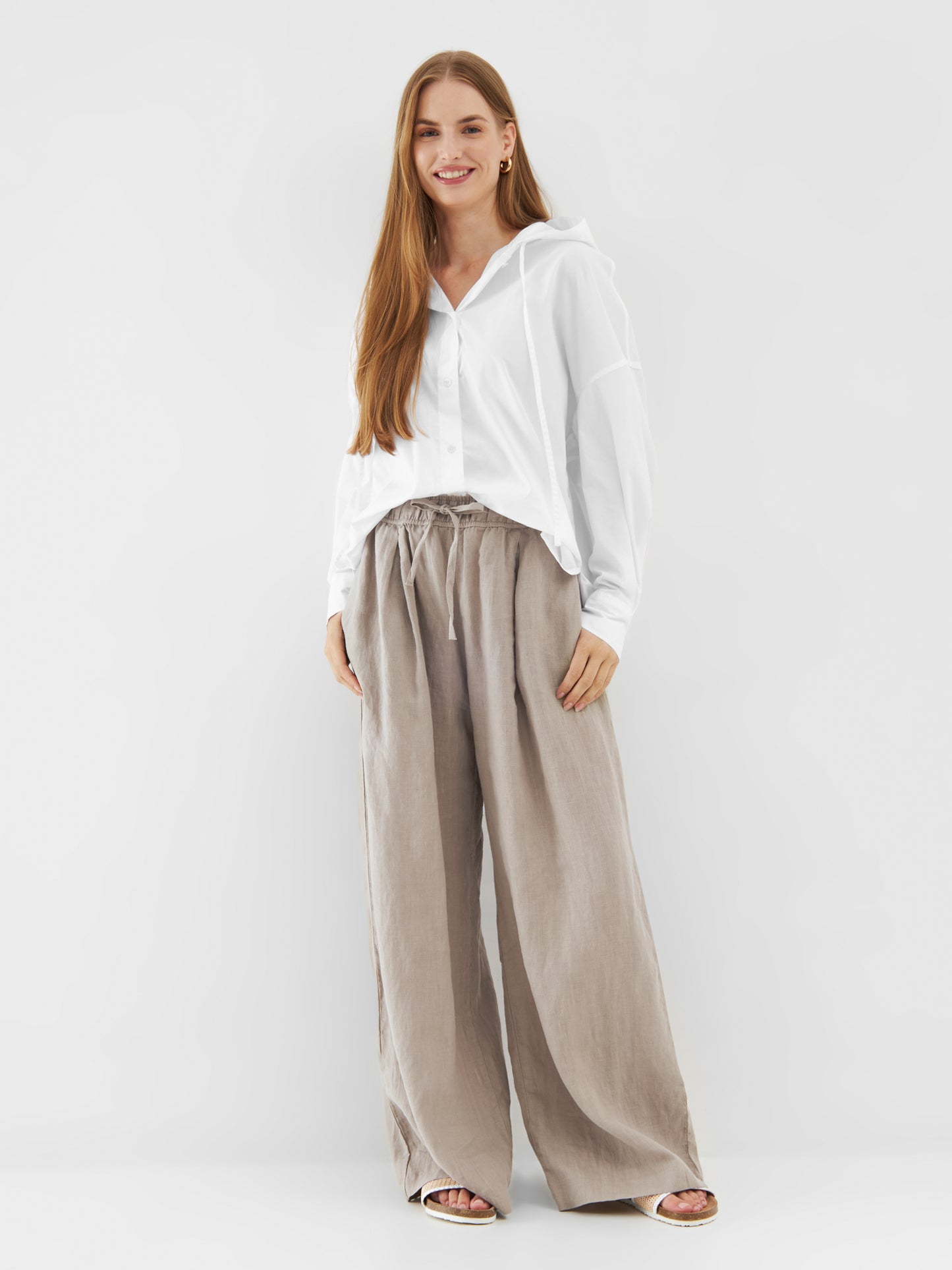 Eine Frau mit langen braunen Haaren lächelt, sie trägt einen Bluse Hoodie White im Stil einer übergroßen Bluse und eine weite beigefarbene Hose mit weitem Bein. Sie steht vor einem schlichten weißen Hintergrund, die Hände locker an der Seite.