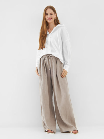 Eine Frau mit langen braunen Haaren lächelt, sie trägt einen Bluse Hoodie White im Stil einer übergroßen Bluse und eine weite beigefarbene Hose mit weitem Bein. Sie steht vor einem schlichten weißen Hintergrund, die Hände locker an der Seite.