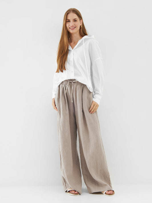 Eine Frau mit langen braunen Haaren lächelt, sie trägt einen Bluse Hoodie White im Stil einer übergroßen Bluse und eine weite beigefarbene Hose mit weitem Bein. Sie steht vor einem schlichten weißen Hintergrund, die Hände locker an der Seite.