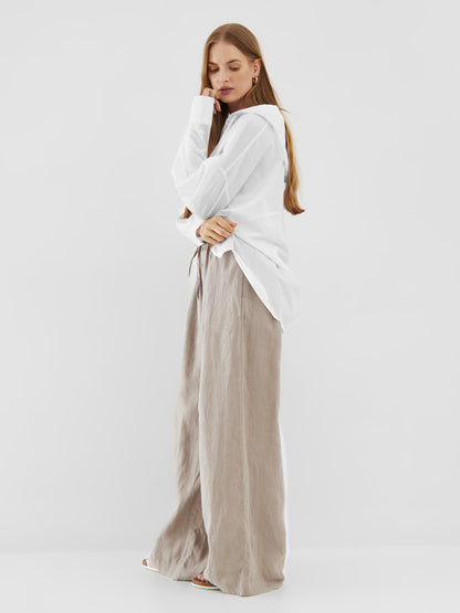 Eine Frau mit langen Haaren steht seitlich vor einem weißen Hintergrund. Sie trägt einen übergroßen Bluse Hoodie White und eine beigefarbene Leinenhose mit weiten Beinen. Sie berührt mit ihrer Hand sanft ihr Kinn.