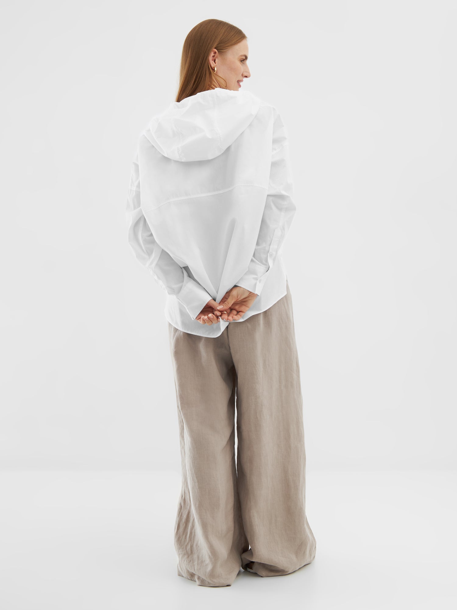 Eine Frau mit glattem, langem Haar steht in einem lockeren Bluse Hoodie White und einer beigefarbenen Hose mit weiten Beinen mit dem Gesicht nach hinten, die Hände hinter dem Rücken verschränkt, vor einem schlichten weißen Hintergrund.