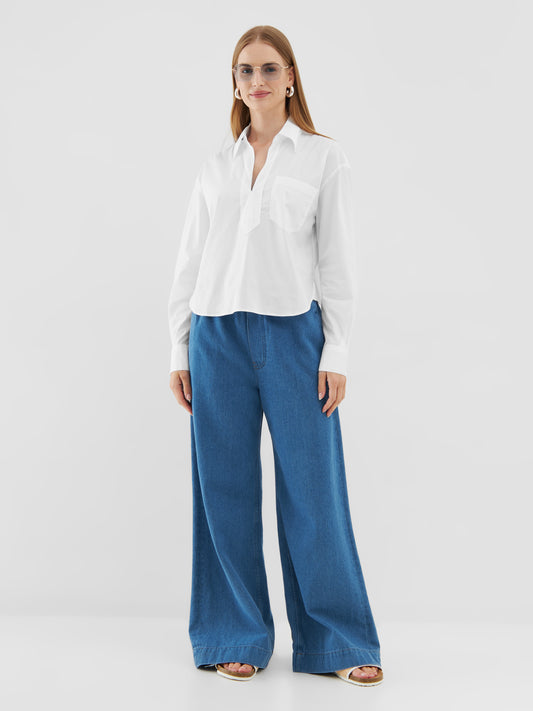 Eine Frau mit glattem Haar trägt ein übergroßes Bluse Polo White, weite Blue Jeans, Sandalen, Ohrringe und eine Sonnenbrille, während sie vor einem schlichten weißen Hintergrund steht.