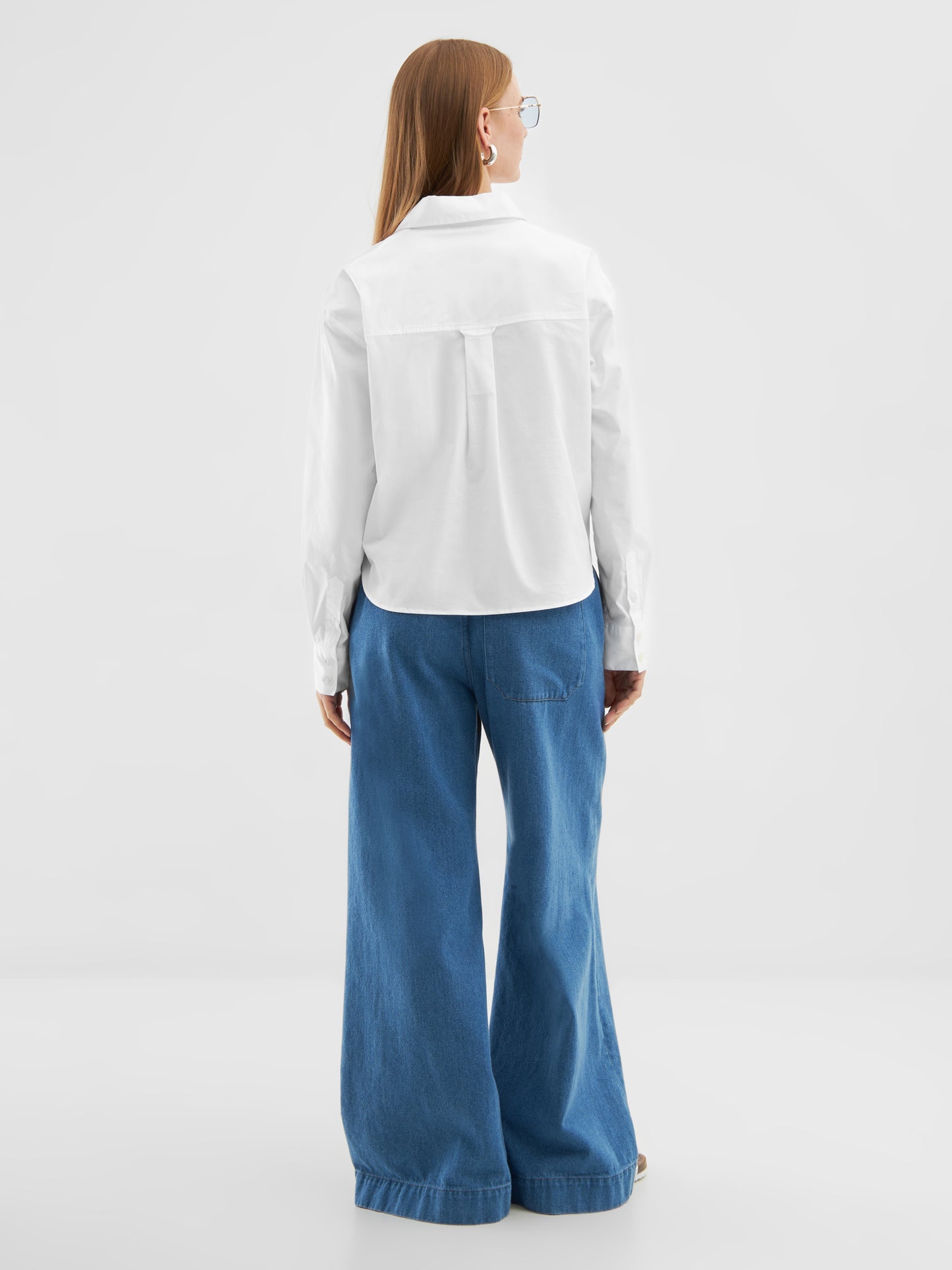 Eine Person mit langem glattem Haar, einer Brille, einem weißen Bluse-Polo und einer weiten blauen Jeanshose steht mit dem Gesicht zur Seite vor einem schlichten hellen Hintergrund.