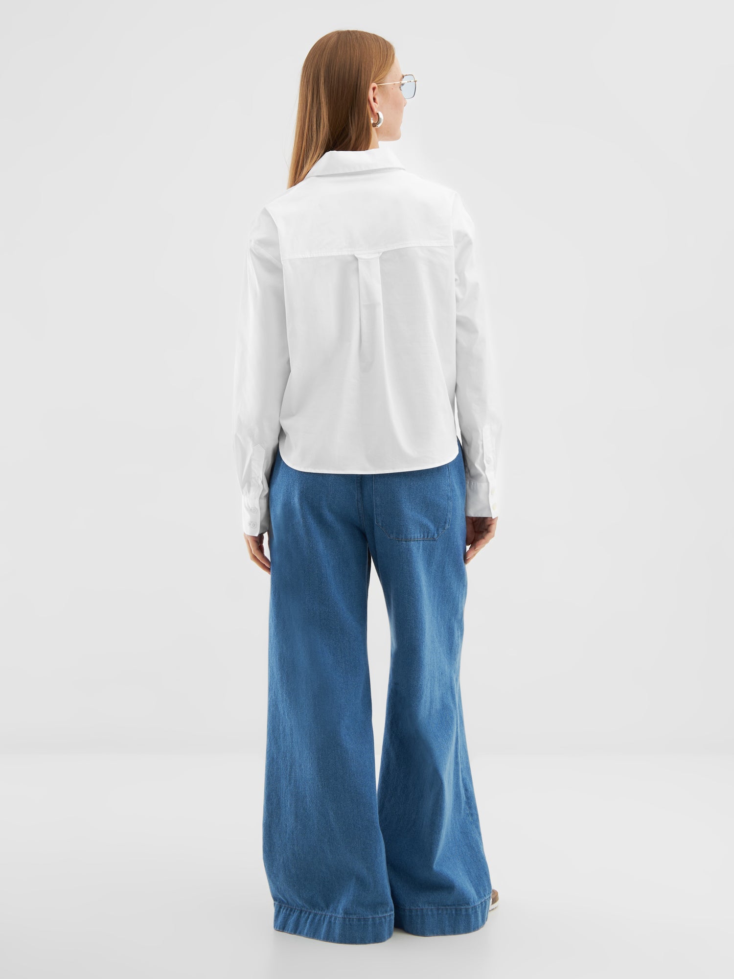 Eine Person mit langem glattem Haar, einer Brille, einem weißen Bluse-Polo und einer weiten blauen Jeanshose steht mit dem Gesicht zur Seite vor einem schlichten hellen Hintergrund.