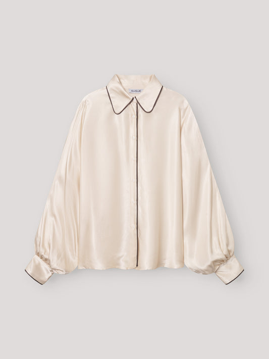 Die cremefarbene Bluse Antonia PJ Satin hat lange, weite Ärmel und schwarze Paspeln am spitzen Kragen, der Knopfleiste und den Manschetten. Diese elegante Damenbluse ist flach auf einem hellgrauen Hintergrund ausgestellt.