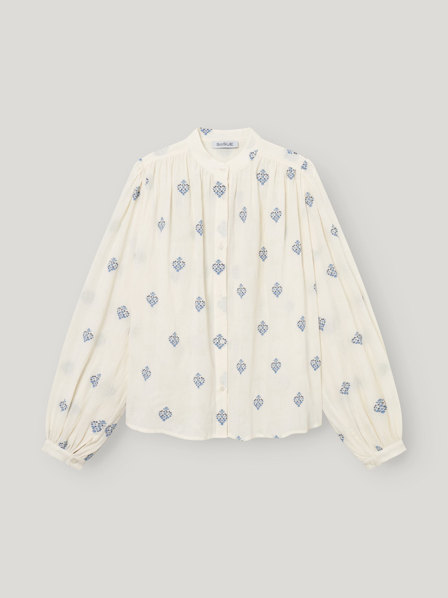 Die Bluse Embroidery ist eine weiße Damenbluse mit langen Ärmeln, einem hohen Kragen, plissierter Vorderseite, blauer geometrischer Blumenstickerei, geknöpften Manschetten und einer entspannten Passform für mühelosen Stil.